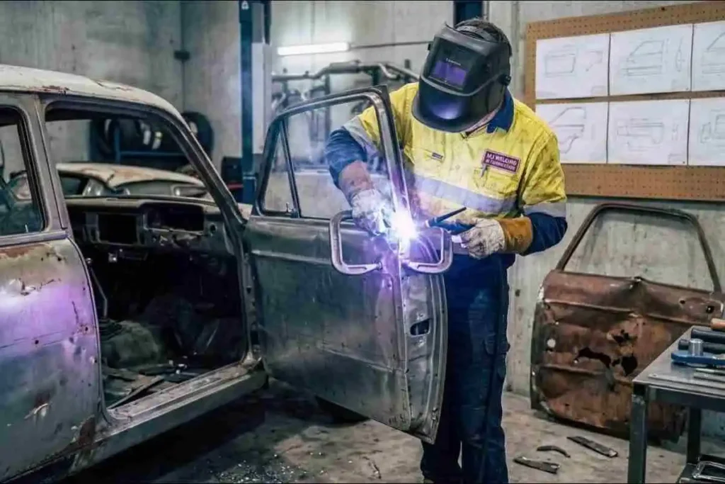 A technician standing beside a stripped vintage car shell, using a C-clamp to hold a new door skin in place while creating a bright white weld arc along the seam.